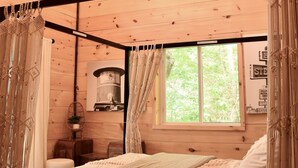 Interno di una baita in legno con un letto, tende in macramè e una finestra vista alberi.