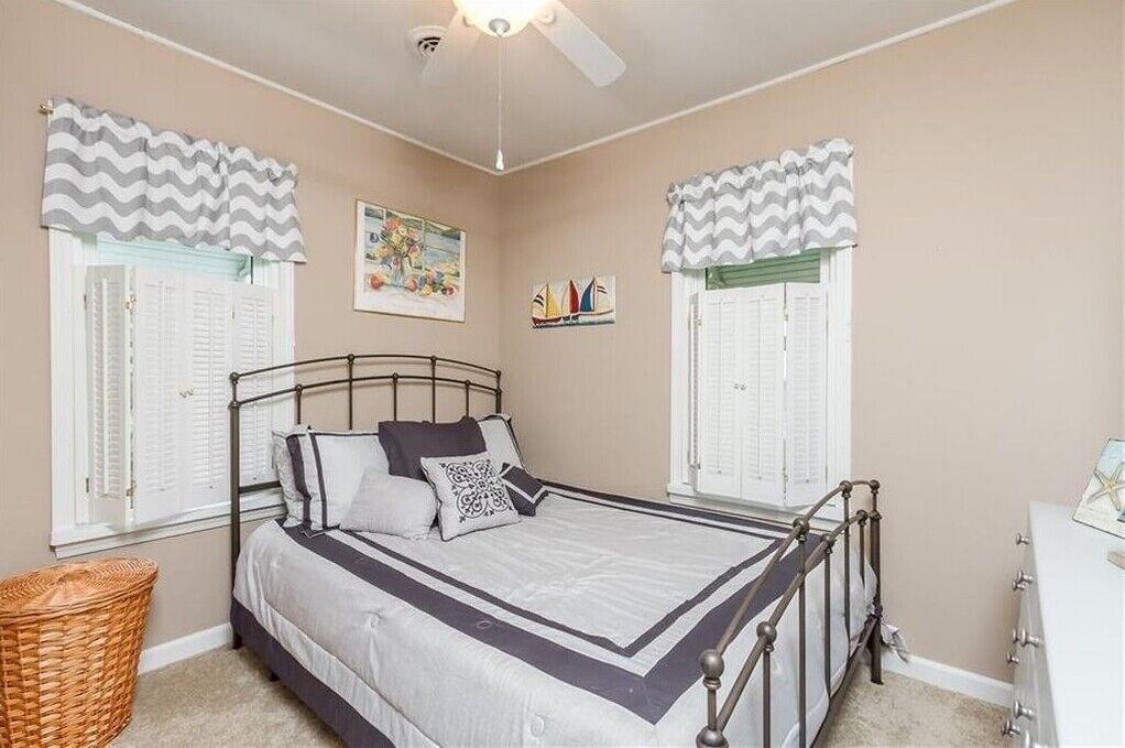 Un dormitorio con una cama, dos ventanas con contraventanas, un ventilador de techo y un estante de pared con objetos decorativos.
