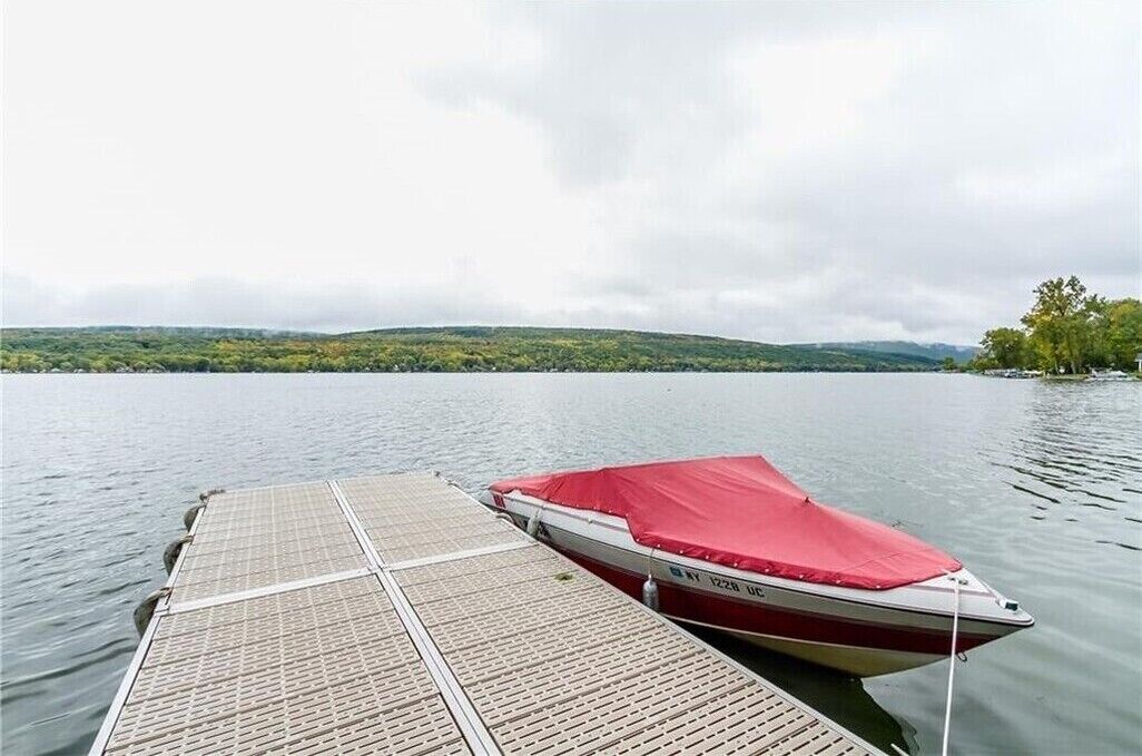 A boat docked at a wooden pier with a red cover, a calm lake, and a hilly landscape in the background.