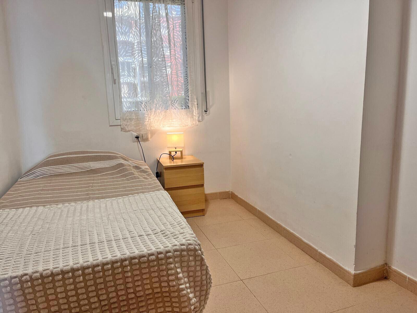 A small, clean room with a bed, a bedside table, and a window with curtains.