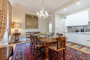Comedor con mesa de madera dispuesta para seis comensales, rodeada de sillas. Un candelabro cuelga del techo y una alfombra estampada cubre el suelo.