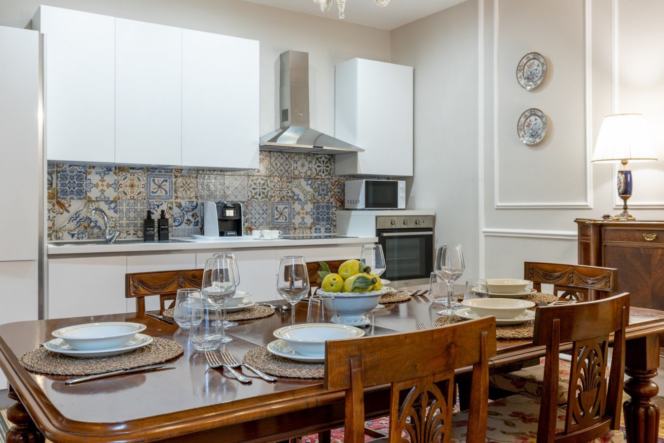 A dining area with a wooden table set for four, a kitchen with white cabinets and a tiled backsplash, and a microwave.