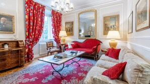 Een kamer met een rode bank, een beige bank, een houten dressoir, een glazen salontafel, een rood gestreept vloerkleed, een kroonluchter en een grote spiegel.