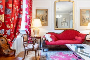 Une pièce avec un canapé rouge, une chaise en bois, une table avec des livres, une lampe et un grand miroir.