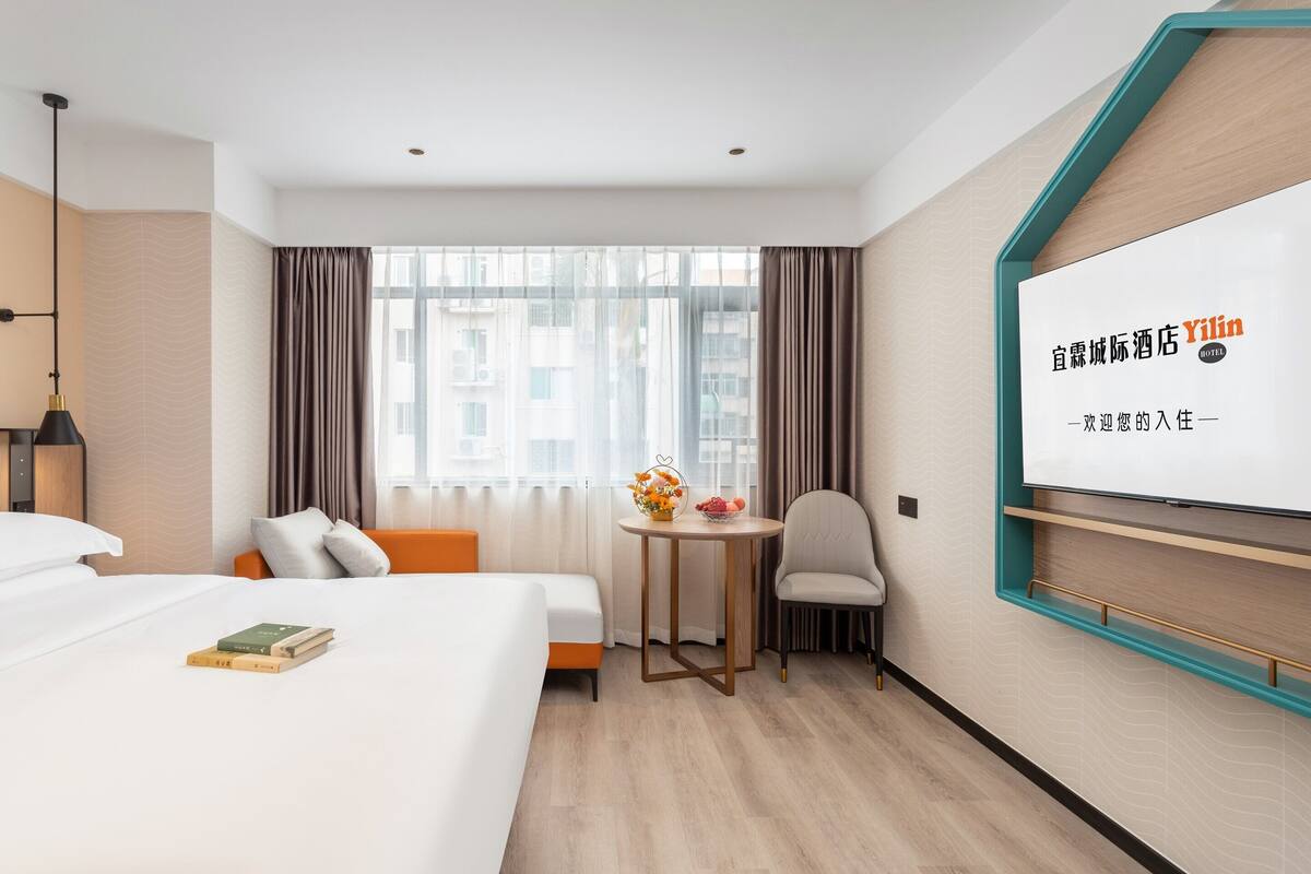 Una habitación de hotel con cama, una mesita de noche con un florero, una silla y un televisor en la pared.