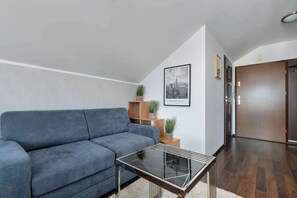 Ein modernes Wohnzimmer mit grauem Sofa, einem Couchtisch mit Glasplatte, Holzregalen mit Pflanzen und einem gerahmten Stadtbild an der Wand.