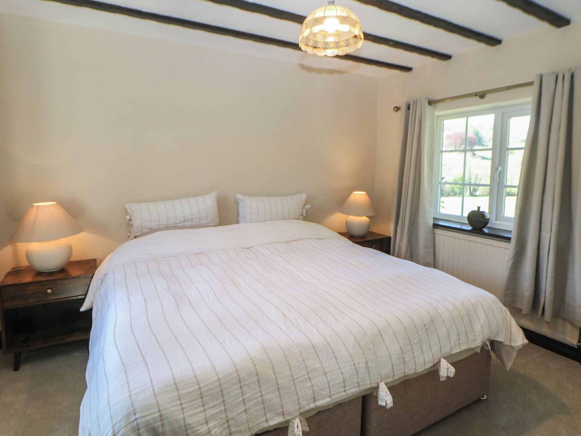 A bedroom with a large bed, two bedside lamps, a window with curtains, and a chandelier.