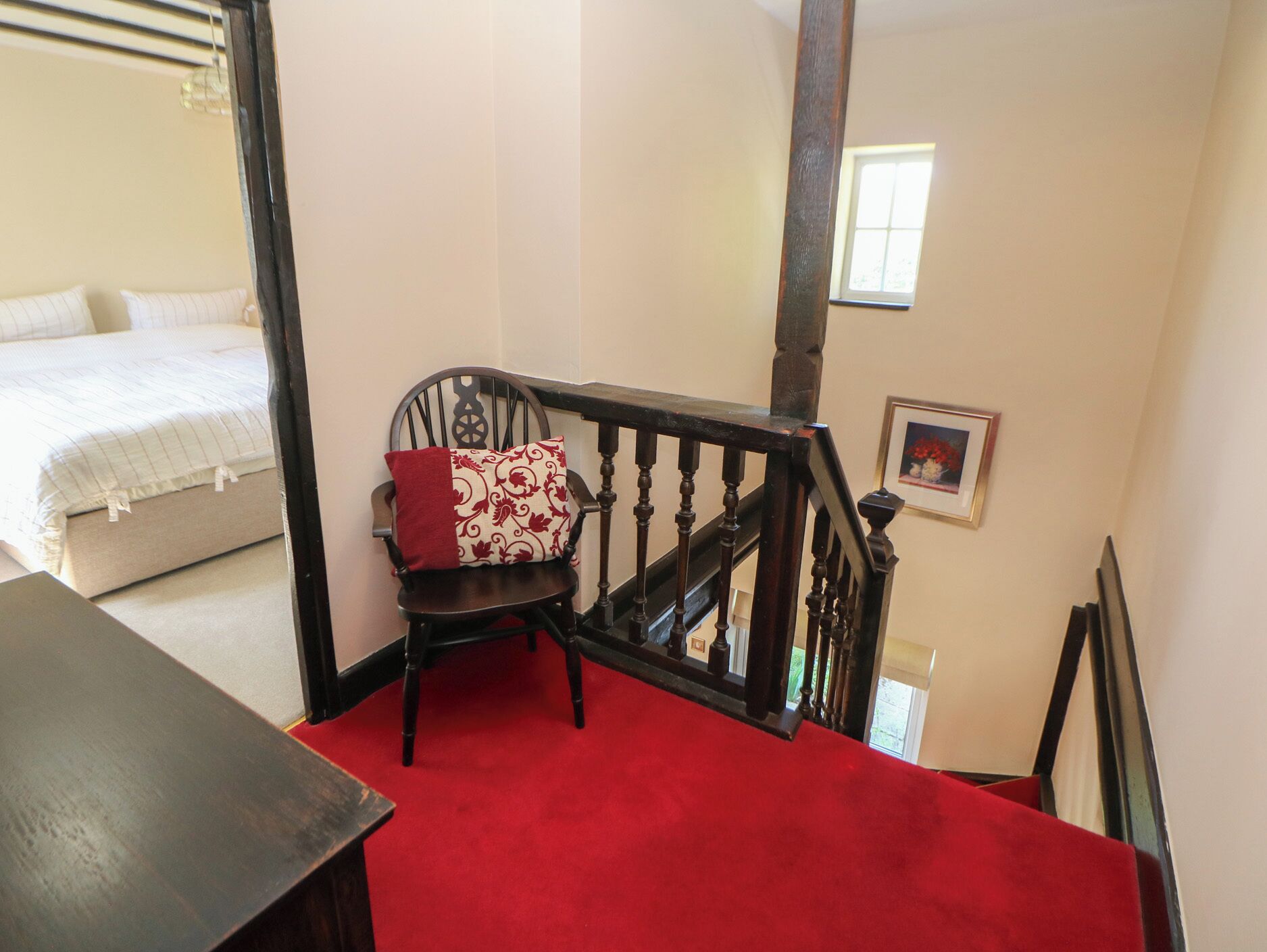 Un dormitorio con cama, una silla con cojín estampado, una escalera con alfombra roja y un cuadro enmarcado en la pared.