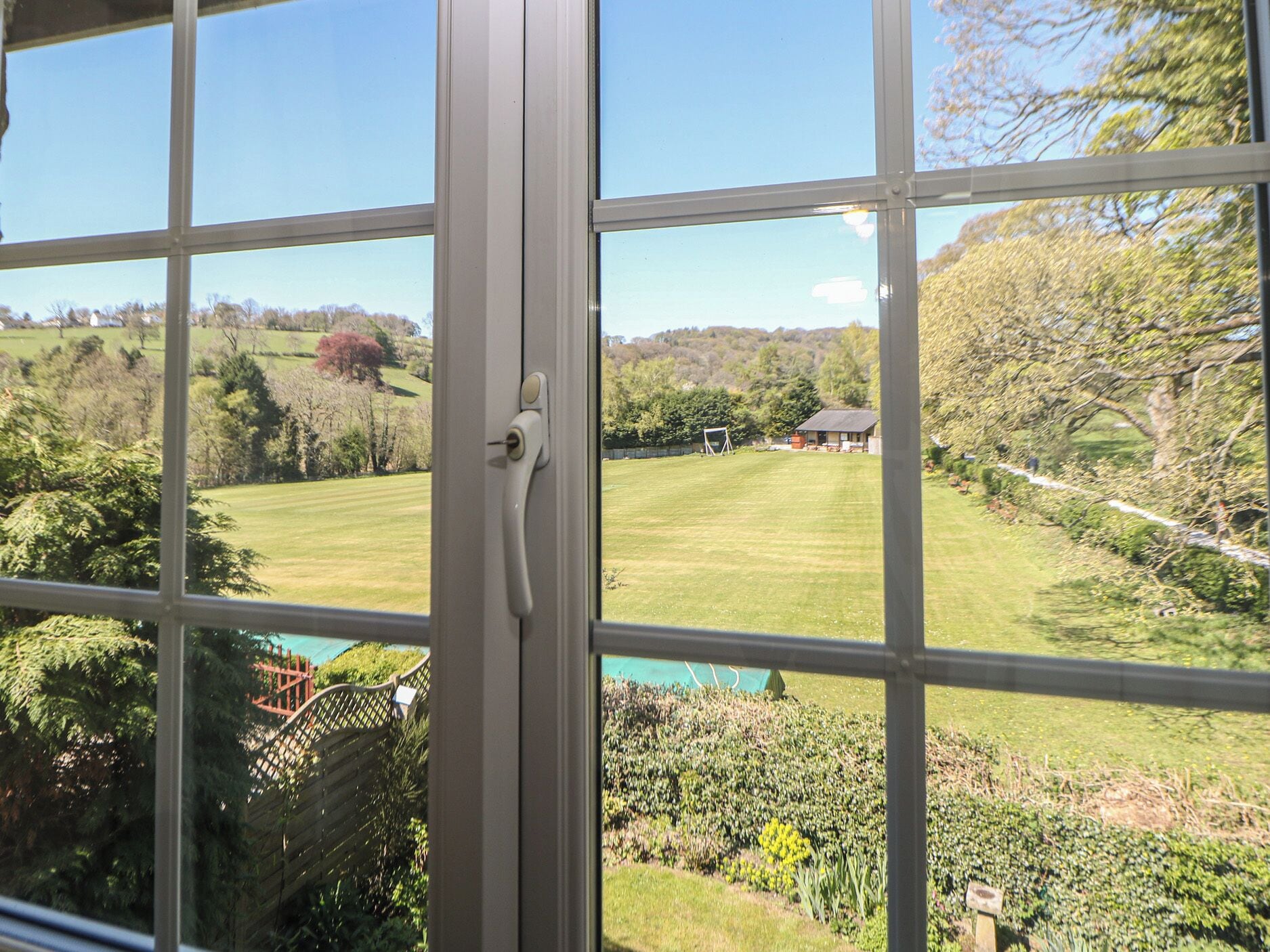 A view through a window with a white frame, a door handle, and a clear view of a green lawn, trees, and a distant building.