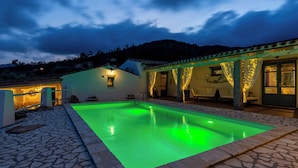 A swimming pool with a stone border, a covered outdoor seating area, and a building with a tiled roof. - BeLocations - Villa VERDALMA (Sardegna)
