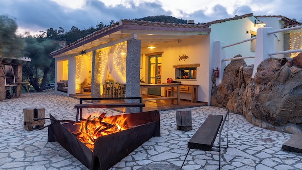 A stone patio with a fire pit, outdoor seating, and a building with a tiled roof and stone walls. - BeLocations - Villa VERDALMA (Sardegna)