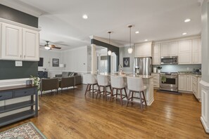 Une cuisine moderne avec des meubles blancs, un îlot central doté de tabourets, un four encastré et un ventilateur de plafond.