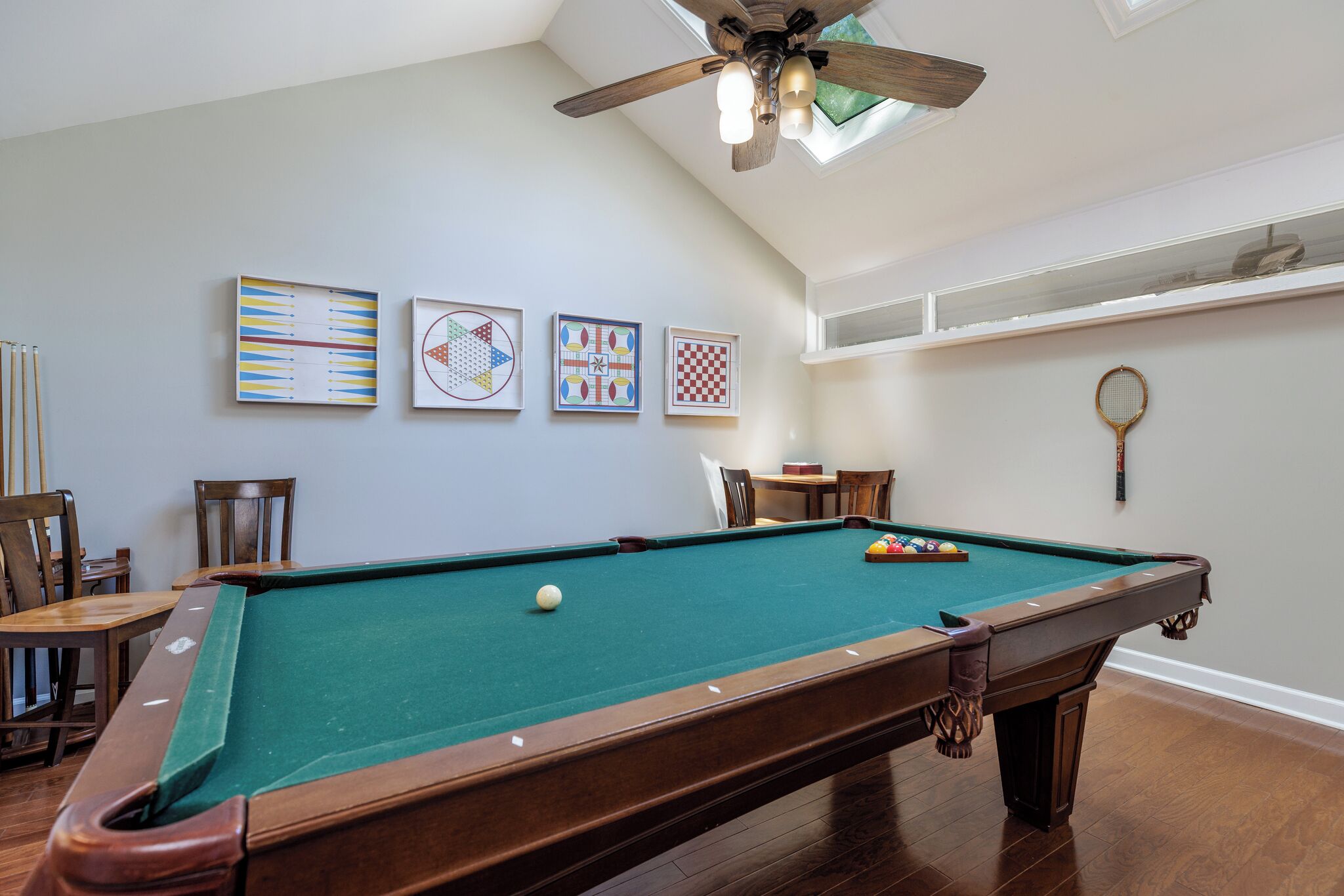 A pool table with a ceiling fan, wooden chairs, and framed artwork on the wall.