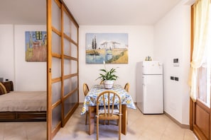 A compact room with a bed, a dining table with chairs, a refrigerator, and two paintings on the walls. - This cozy ground-floor apartment, ideal for 3 people, is perfect for a relaxing stay in Siena. The apartment overlooks the shared garden, where you can enjoy outdoor dining with a table, armchairs, and chairs with an umbrella. The location is convenient  (Siena)