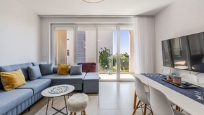 A modern living room with a sofa, a round coffee table, a small stool, a dining area with a table and chairs, and a TV mounted on the wall. - Golden Hour Balcony Apartment | Pool & Beach 100m by PikHost (Tale)