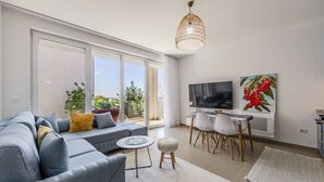A modern living room with a sofa, a dining table with chairs, a TV, and a large window with a view of greenery. - Golden Hour Balcony Apartment | Pool & Beach 100m by PikHost (Tale)