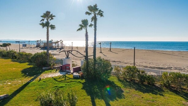 A beach view with palm trees, a fenced area, and a clear sky. - Golden Hour Balcony Apartment | Pool & Beach 100m by PikHost (Tale)