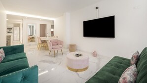 A modern living room with a green sofa, a round ottoman, a pink chair, and a flat-screen TV mounted on the wall.