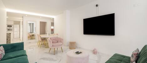 A modern living room with a green sofa, a round ottoman, a pink chair, and a flat-screen TV mounted on the wall.