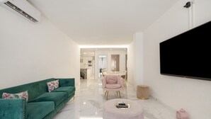 A modern living room with a teal sofa, a pink ottoman, a flat-screen TV, and a dining area in the background.