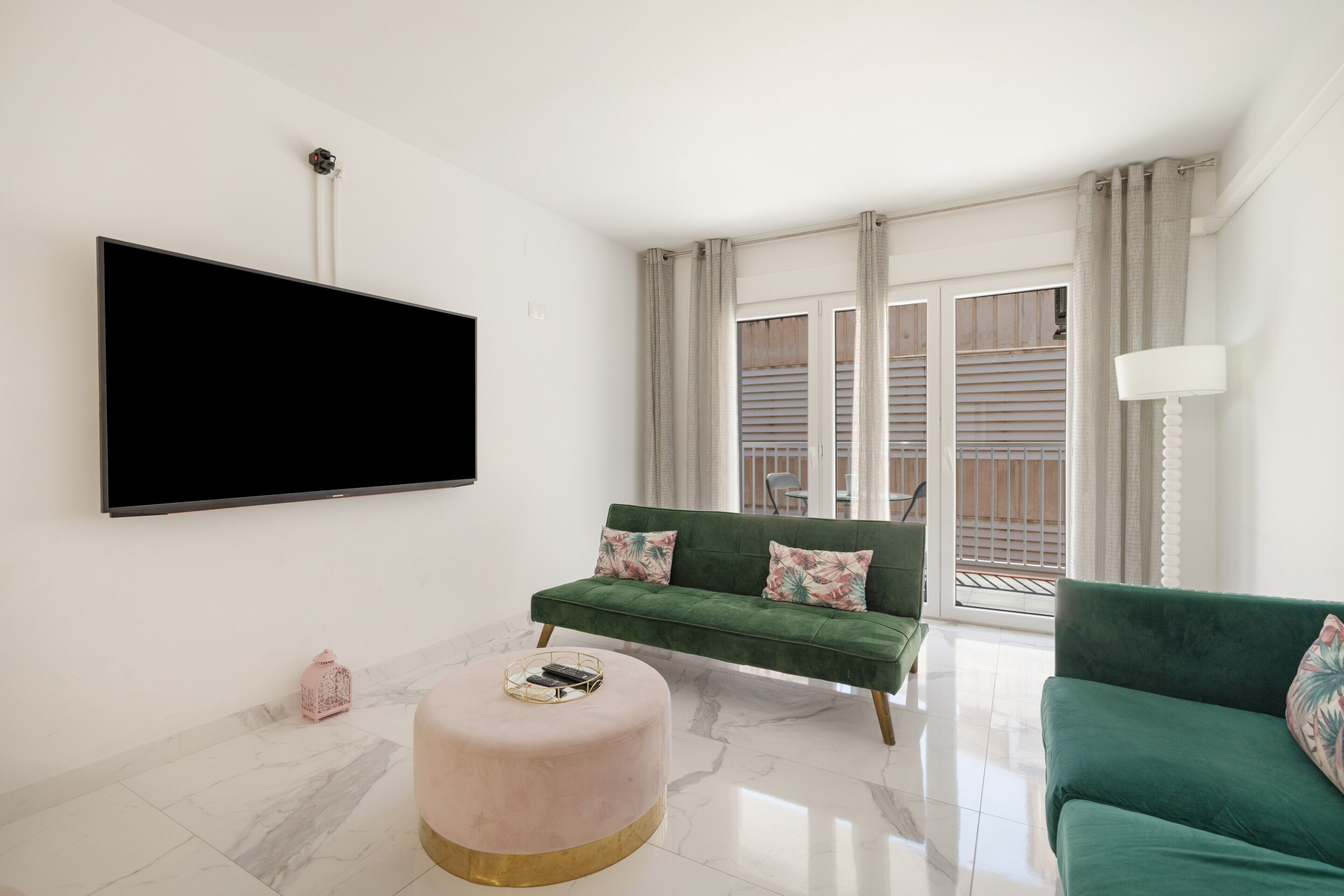 Un salon équipé d’un téléviseur à écran plat, de deux canapés verts, d’un pouf rond et d’une grande fenêtre avec des stores.