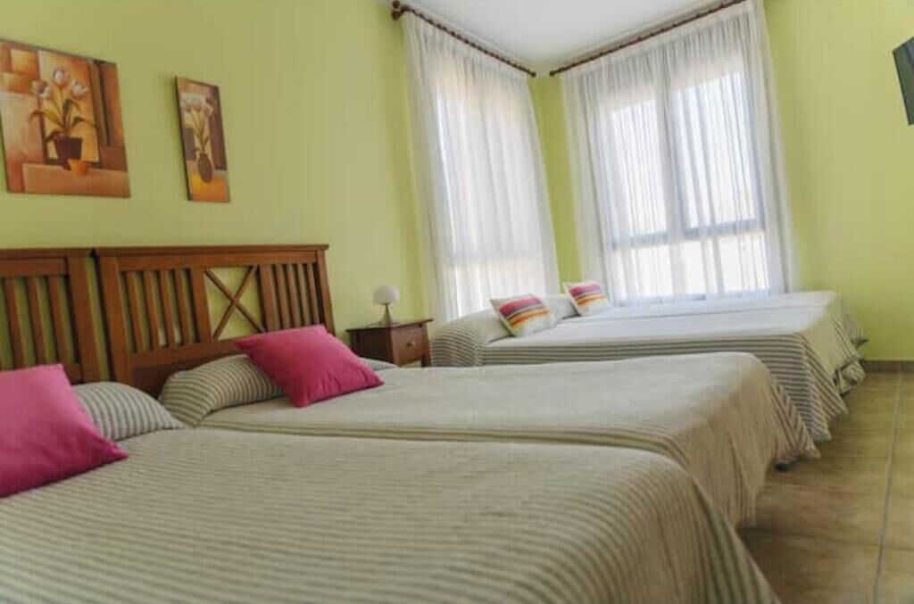 Habitación de hotel con dos camas, cabecera de madera y dos ventanas con cortinas traslúcidas.