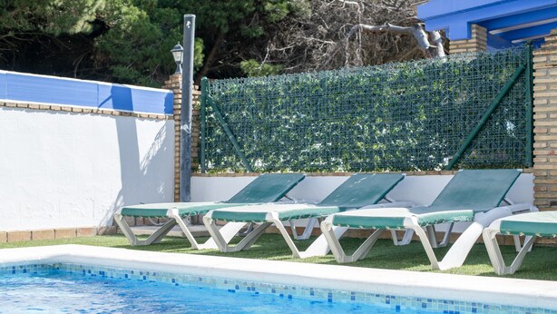 En swimmingpool med liggestole, en murstensvæg og et grønt hegn.