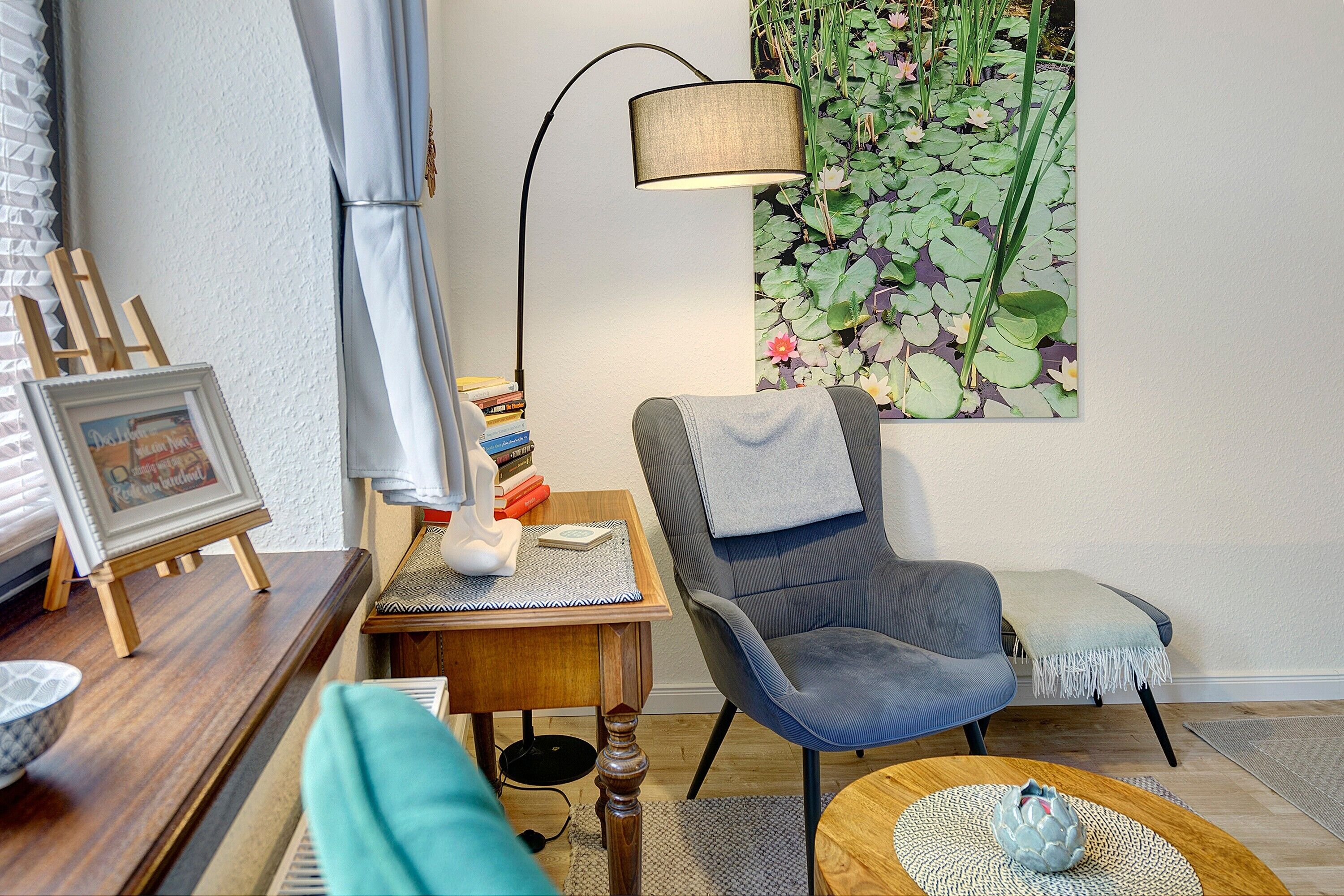 Ett rum med en stol, ett bord, en lampa, en liten tavla och en vägg med blommig tapet.