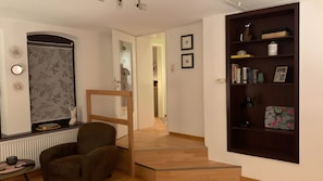 Un intérieur moderne avec un escalier en bois, un coin salon comprenant un fauteuil, une bibliothèque et une porte donnant sur une autre pièce.