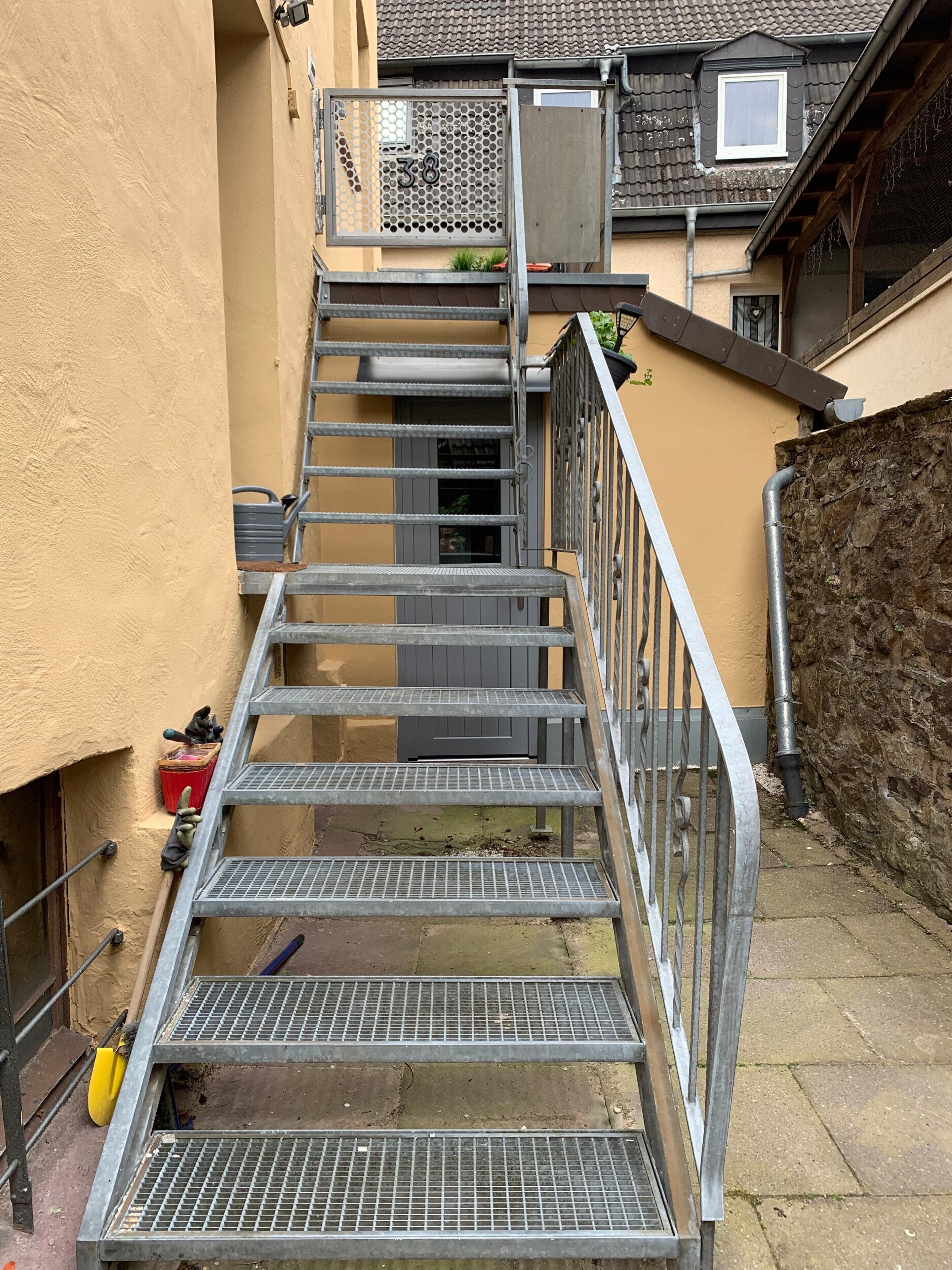 Una escalera metálica conduce a la entrada de un edificio, con un letrero con el número 38, una caja de herramientas roja y una pala amarilla en el suelo.