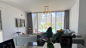 En moderne stue med sofa, fjernsyn, et bord med en plante og store vinduer med udsigt til bygninger og en pool.