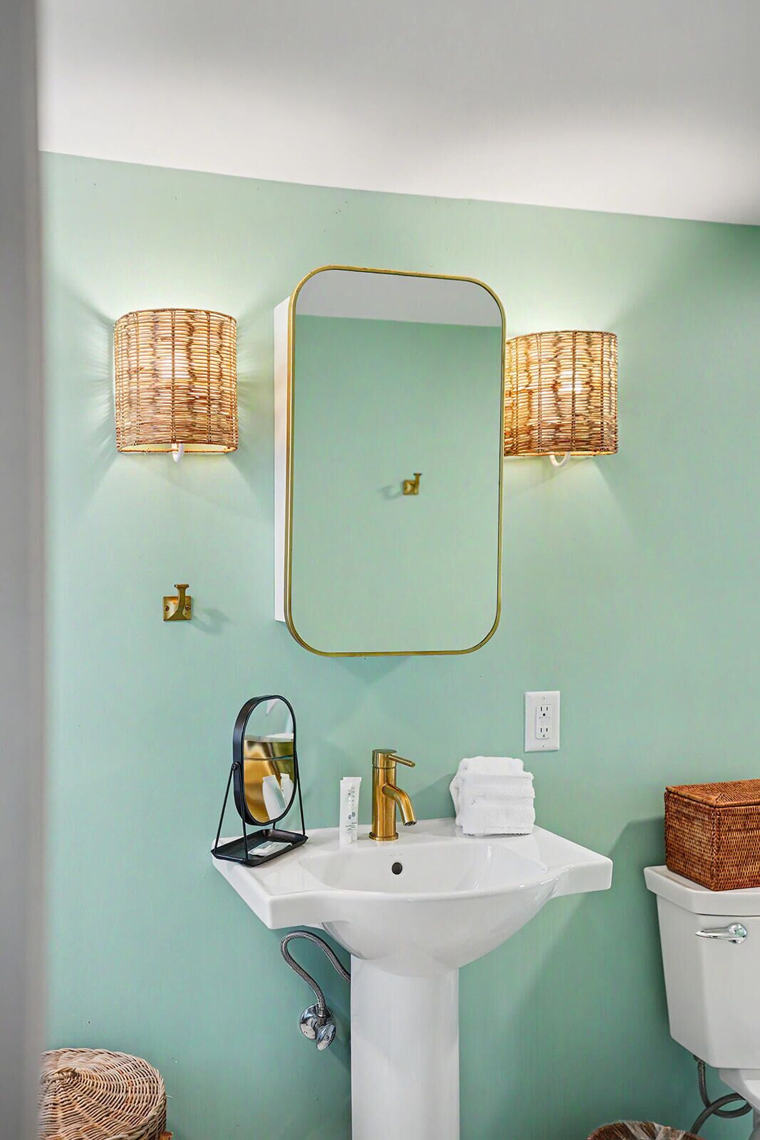 Un baño con lavamanos blanco, grifo dorado y un espejo grande con marco dorado. Hay dos apliques de pared con pantallas de mimbre.