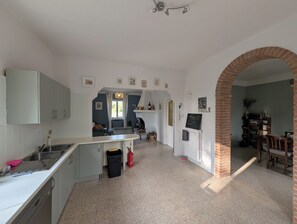 Eine Küche mit weißen Schränken, einer Spüle und einem Feuerlöscher. Es gibt eine Essecke mit Tisch und Stühlen sowie einen Ziegelbogen.