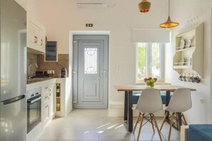 A modern kitchen with white cabinets, a built-in oven, a dining area with a table and chairs, and a window with a view. - Joy’s Apartment (Milos)