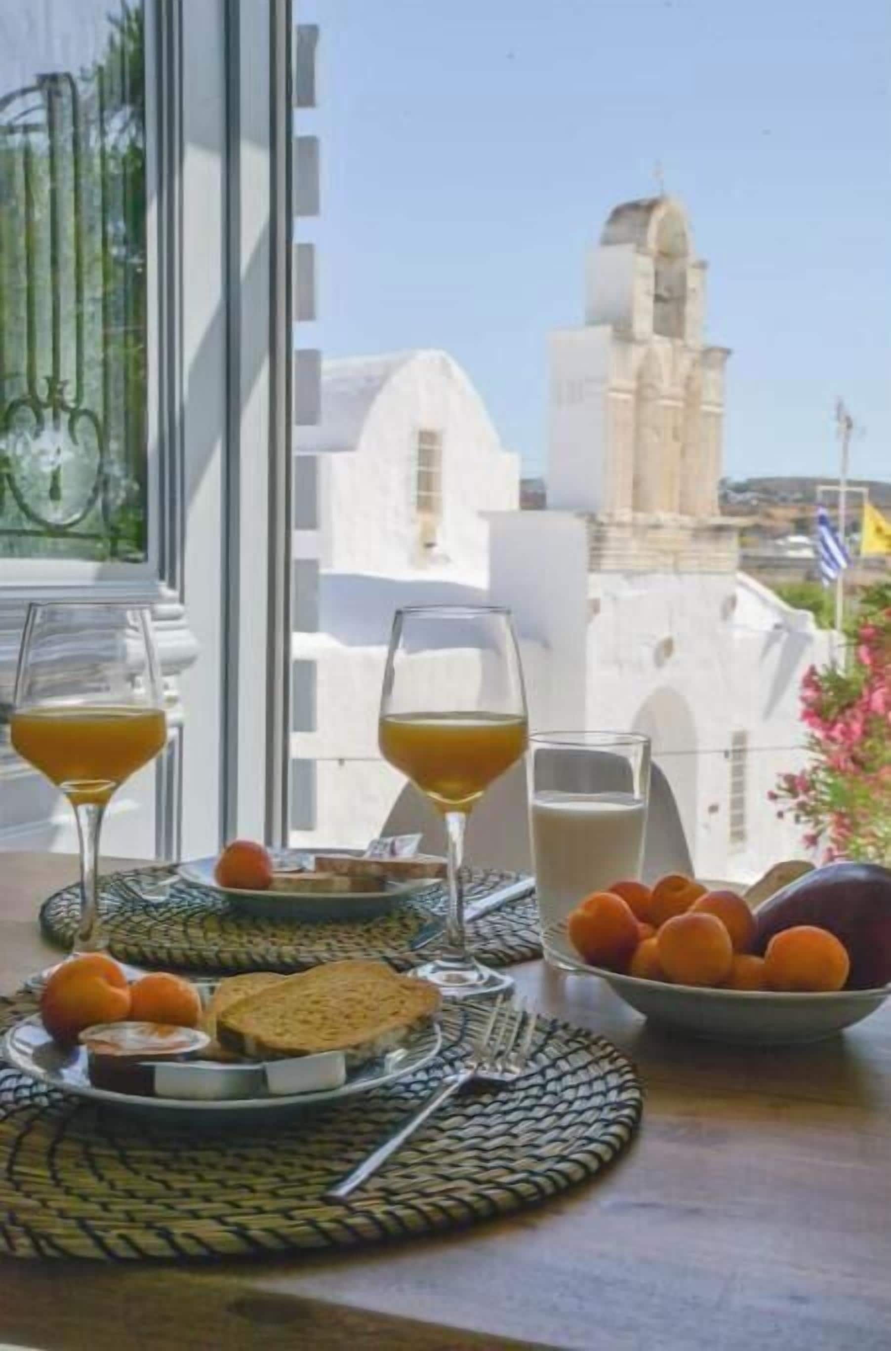 A breakfast table with glasses of orange juice and milk, a plate with bread and fruit, and a bowl of oranges.
