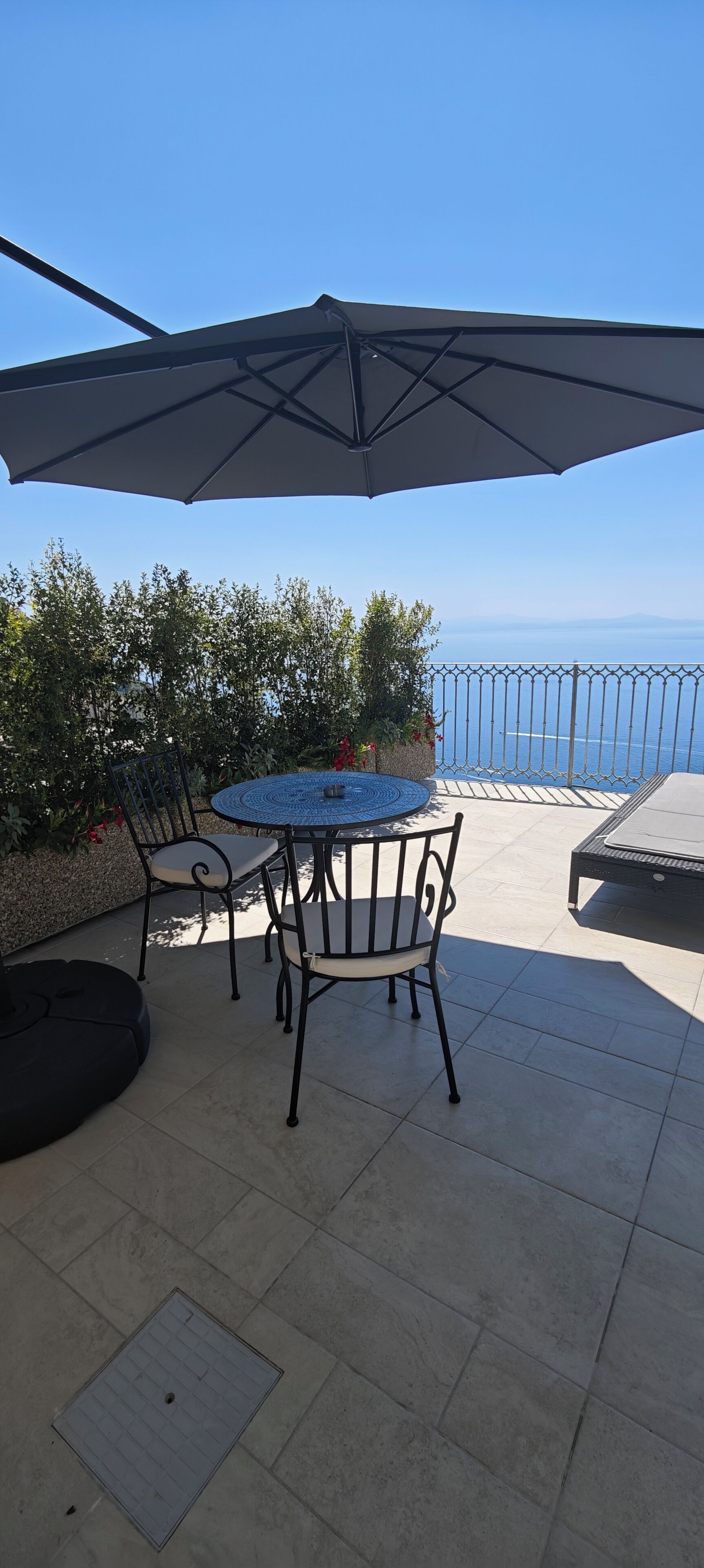 An outdoor patio with a round table and chairs, a large umbrella, and a view of the sea.