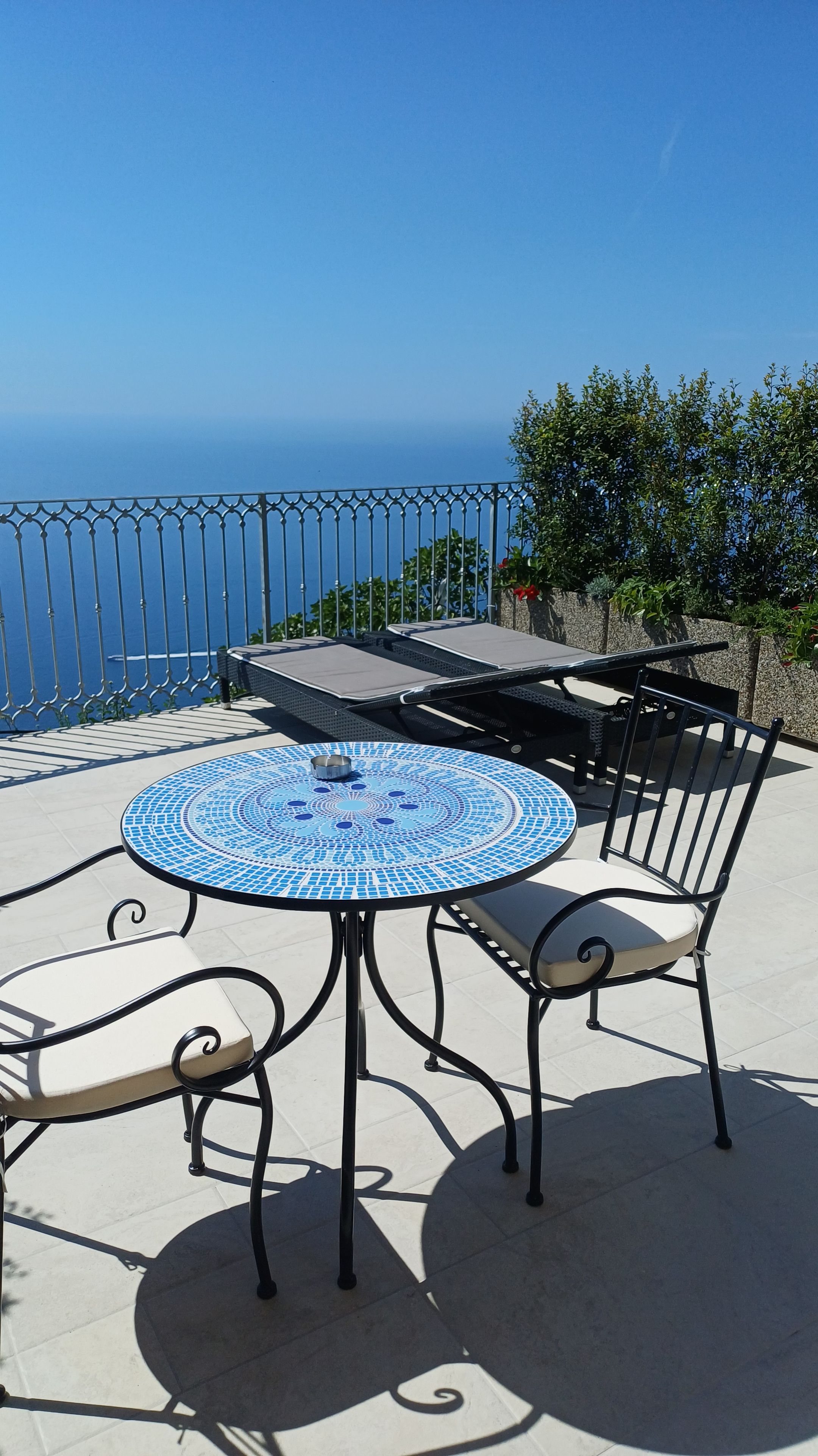 Ein runder Tisch mit blauer Mosaikplatte, zwei Stühle und Blick aufs Meer.