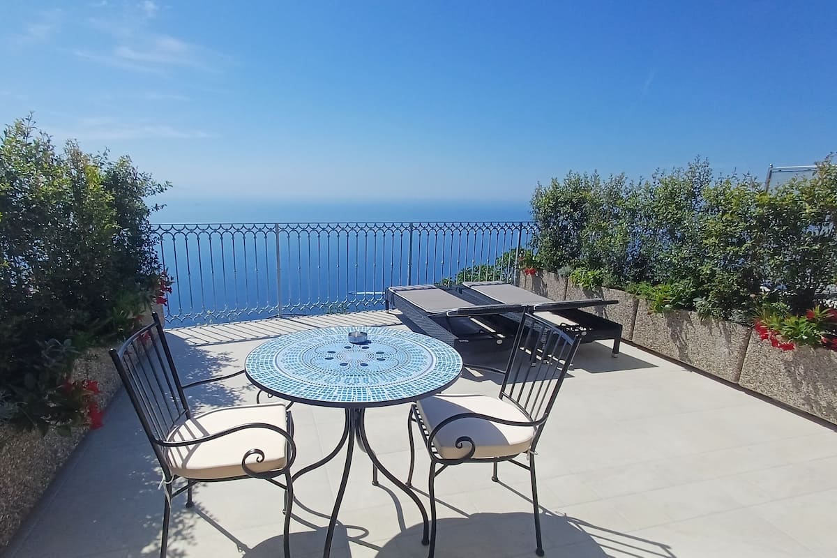 Une terrasse avec une table ronde et des chaises, une vue sur la mer et une rambarde en métal.