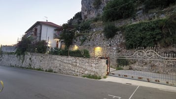 A stone wall with a metal gate, a paved road, and a building with a red roof.