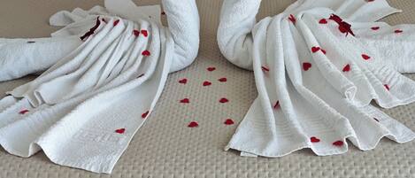 A bed with a headboard, a white bedspread, and two towels arranged in a swan shape with red heart decorations.