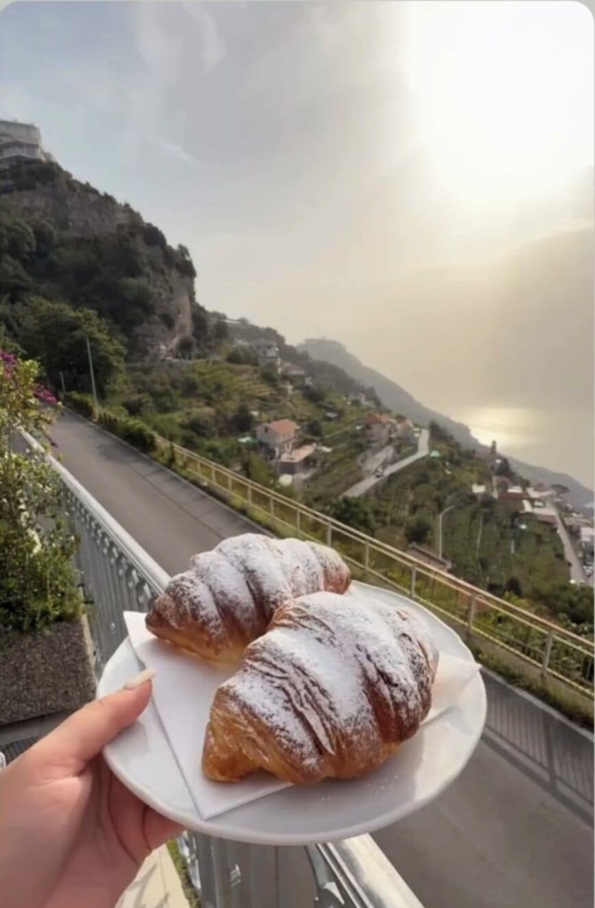 Une main tenant une assiette avec deux pâtisseries, avec en arrière-plan un paysage montagneux, une route et des bâtiments.