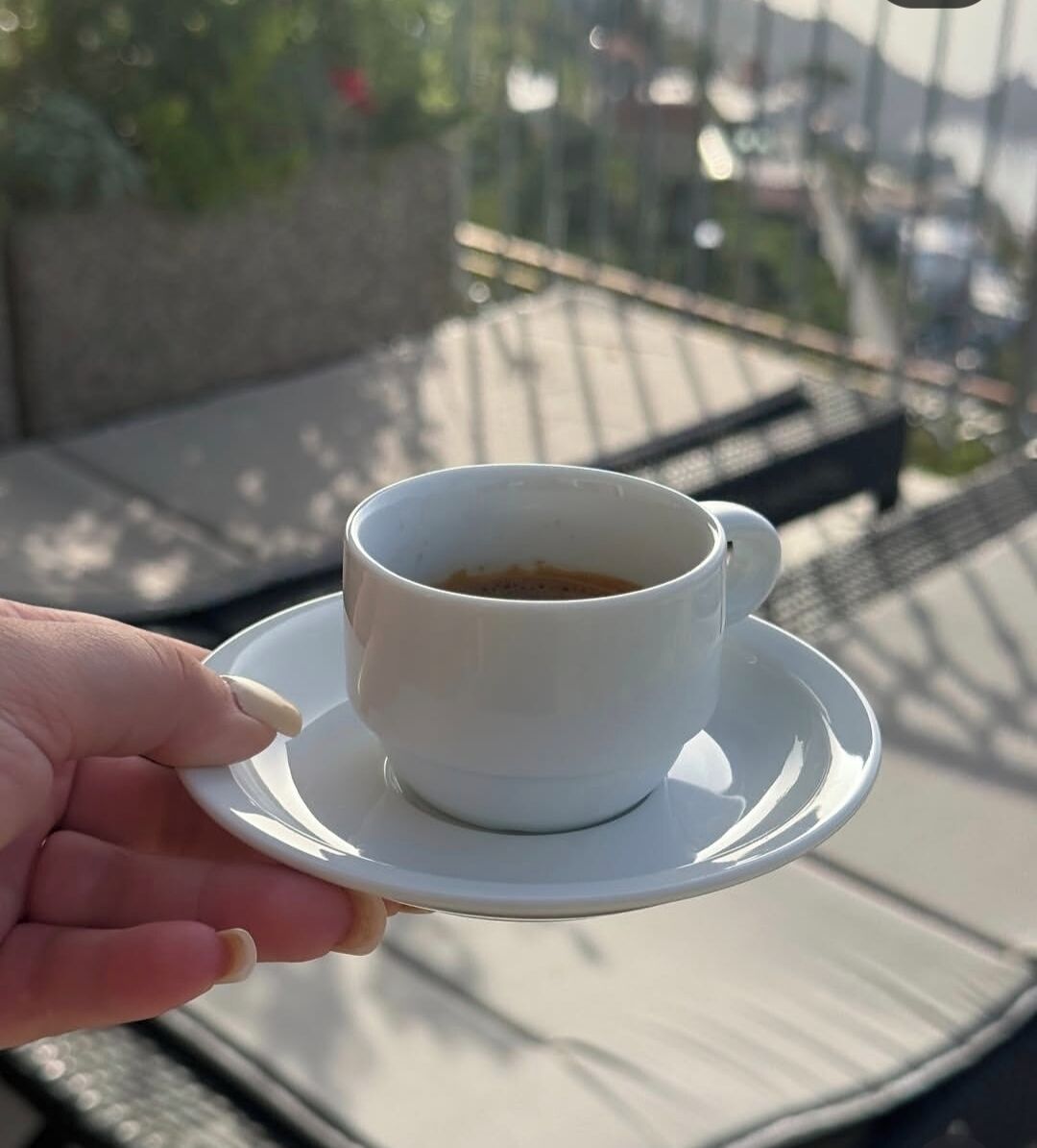 Una mano sostiene una taza blanca de café sobre un platito, con un fondo exterior desenfocado.
