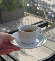 Una mano sostiene una taza blanca de café sobre un platito, con un fondo exterior desenfocado.