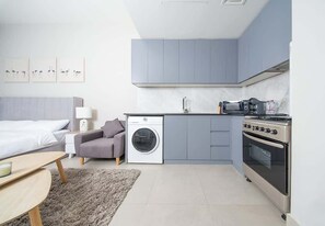 A compact hotel room with a bed, a sofa, a washing machine, a kitchenette with a stove, and a small table. - Studio w/ Pool & Gym at JVC Living Garden (Dubai)