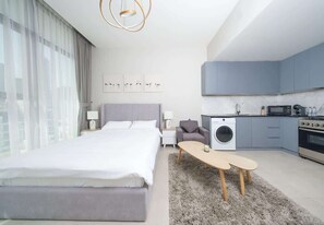 A modern hotel room with a large bed, a washing machine, a kitchenette, and a view of the city. - Studio w/ Pool & Gym at JVC Living Garden (Dubai)