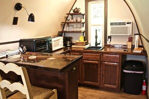 A compact kitchen with a microwave, toaster, and sink. There is a shelf with cups and a bag of groceries.