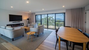 Uma sala de estar moderna com televisão de tela plana, sofás cinzas, mesa de jantar de madeira e uma grande janela com vista.