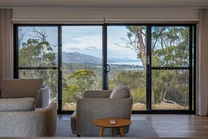 A room with a large sliding glass door, a sofa, a small round table, and a view of a natural landscape. - Freycinet Residences (Coles Bay)