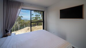 Quarto com cama grande, janela com cortinas, televisão na parede e vista para árvores e colinas.