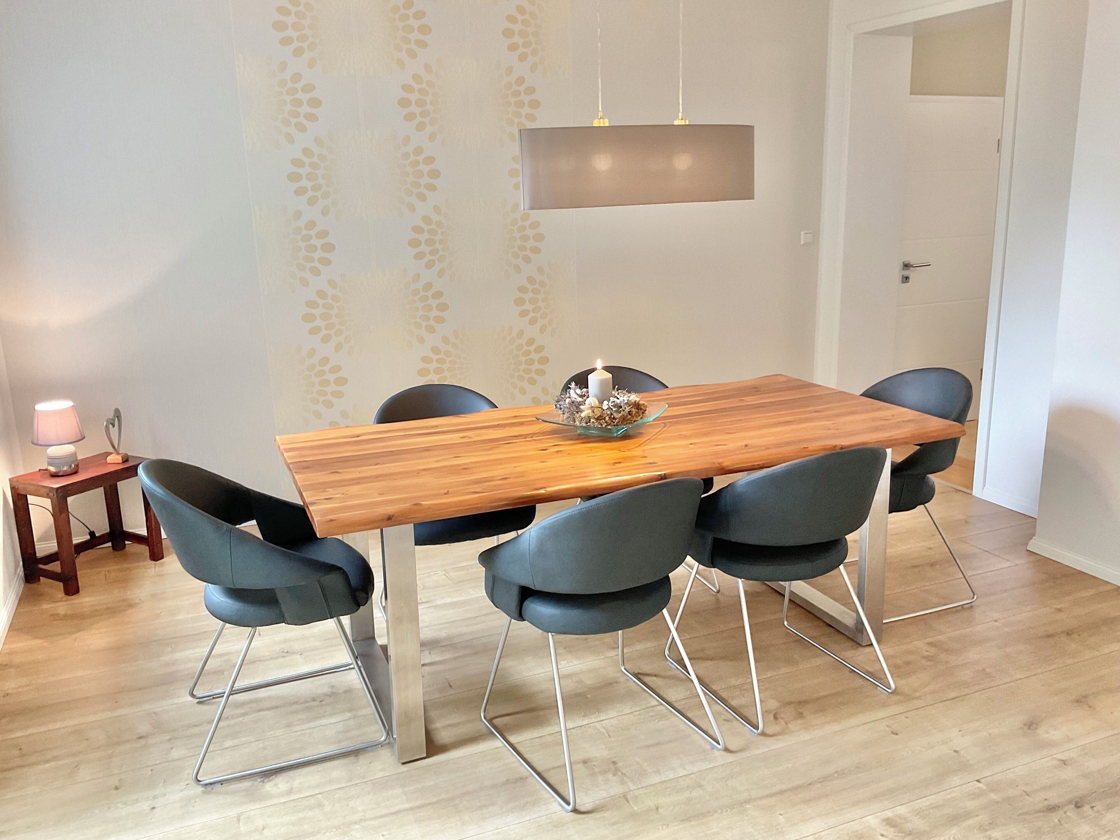 A dining area with a wooden table, six chairs, a hanging light fixture, and a patterned wall.